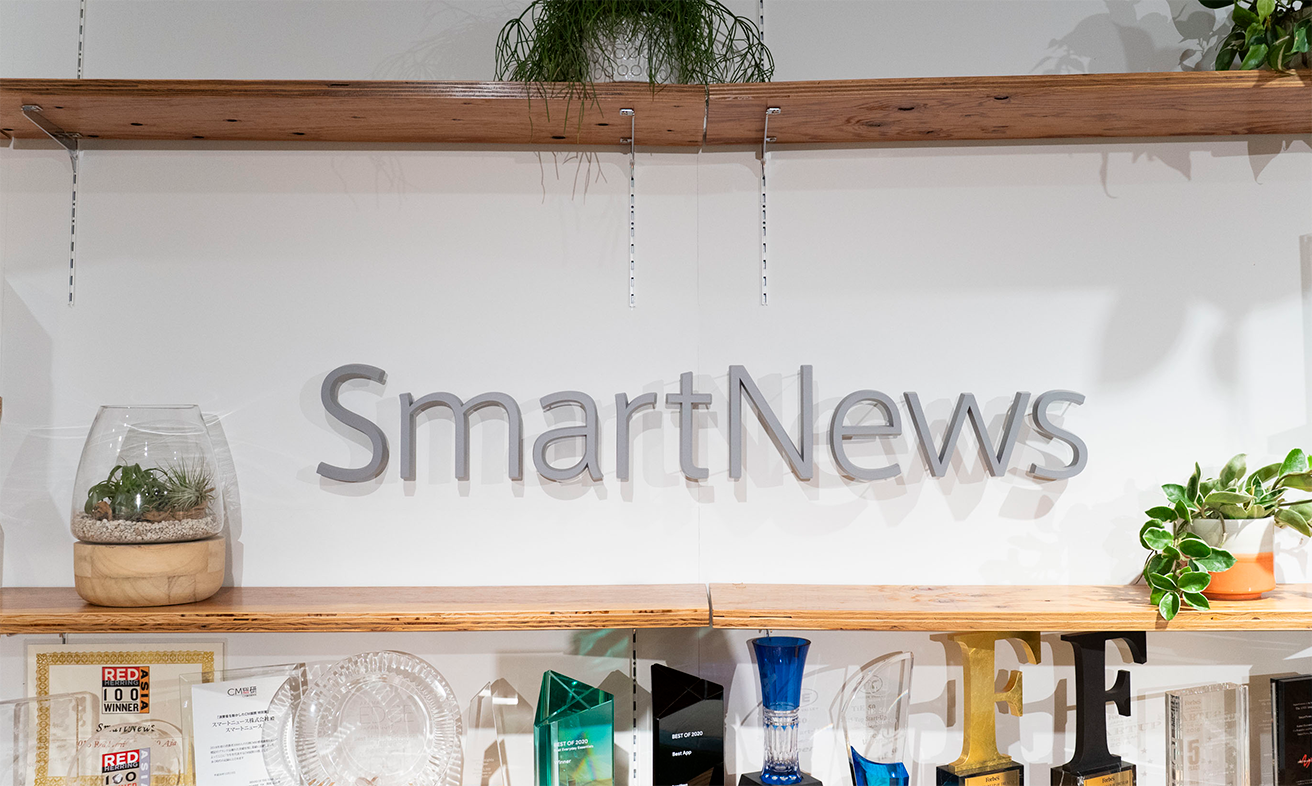 写真：SmartNews社内の様子。白い壁にSmartNewsと書かれた立体的な文字が掲げられている。その下に数々のトロフィーがおかれている。